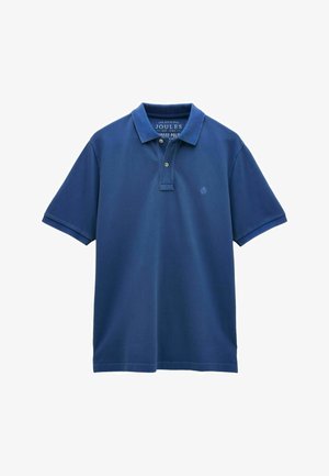 Mann trägt blaues Polohemd, beige Shorts und gemusterte Slip-On-Schuhe, steht vor einer schlichten beigen Wand.