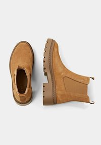 Botas de tobillo de ante marrón con paneles laterales elásticos, suelas de goma marrón gruesas y tiradores para ponerse fácilmente. Textura suave con un brillo sutil.