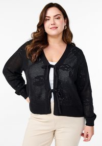 Črn pleten cardigan z V-izrezom, odprto tkanino in sprednjim vezanjem. Ima rebraste manšete in spodnji del ter se dobro poda k beli majici.