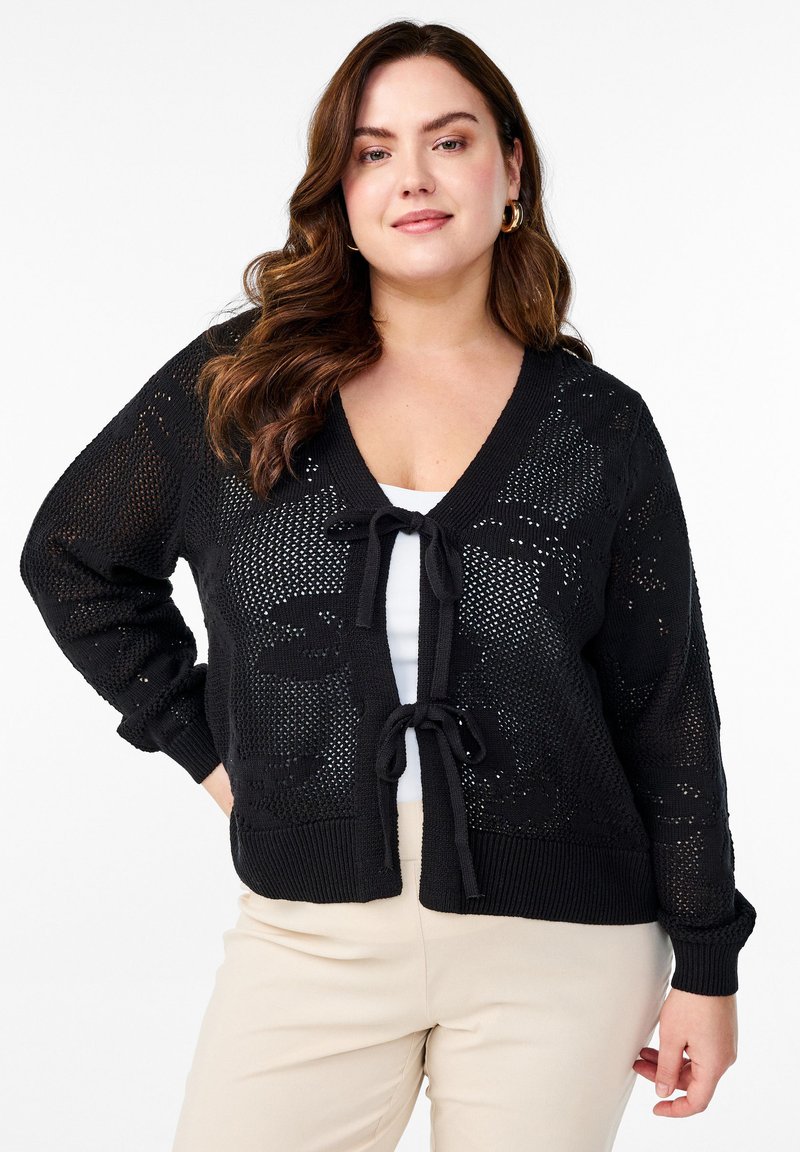 Črn pleten cardigan z V-izrezom, odprto tkanino in sprednjim vezanjem. Ima rebraste manšete in spodnji del ter se dobro poda k beli majici.