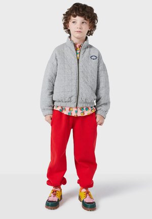 Grau gesteppte Bomberjacke mit Reißverschluss, rote lässige Hose und mehrfarbige Schuhe mit strukturierten Akzenten. Darunter ist ein gemustertes Hemd geschichtet.