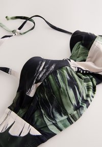 Haut de bikini avec liens ajustables, présentant un imprimé abstrait de feuilles en noir, blanc et vert sur un tissu lisse, posé à plat sur une surface blanche.