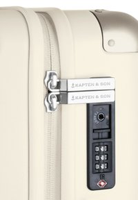 Hartschalentrolley in mattem Creme, ausgestattet mit zwei silbernen Reißverschlüssen, einem integrierten Zahlenschloss und dem Branding "Kapten & Son".