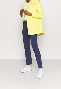 Gul vattentät jacka med huva, fickor med dragkedja och tryckknappar, i kombination med navy slim-fit byxor och vita sportskor.