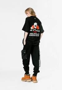 Camiseta negra de corte oversize con estampado gráfico, combinada con pantalones cargo negros con bolsillos laterales, y botas marrones con cordones y suela gruesa.