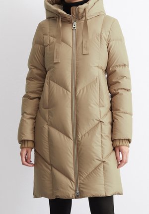 Femme portant un manteau matelassé beige mi-long avec capuche et fermeture éclair à l’avant, associé à un pantalon noir et des poignets tricotés couleur fauve visibles.