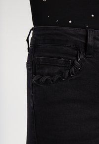 Los jeans de denim negros presentan una cintura alta, costuras sutiles en los bolsillos y un detalle decorativo de cuerda a lo largo del bolsillo lateral.