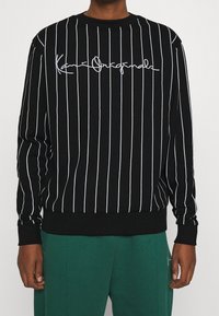 Černá mikina s vertikálními bílými pruhy, na hrudi s bílým nápisem v písmu. Hladké manžety a lem, jemná textura.