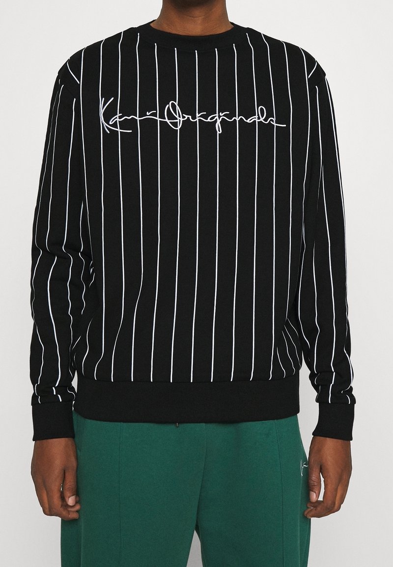 Černá mikina s vertikálními bílými pruhy, na hrudi s bílým nápisem v písmu. Hladké manžety a lem, jemná textura.