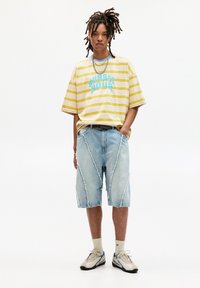 Yellow striped oversized t-shirt with "SUNSET HOUR" printed in blue. Light blue ripped denim shorts with diagonal seam details. Neutral sneakers.
