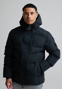 Jeune homme aux cheveux courts portant une veste d'hiver noire matelassée à capuche avec des poches zippées et un logo de la marque sur la manche.