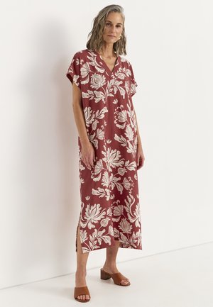 Frau trägt ein lockeres, rostfarbenes Midikleid mit großen weißen Blumenmotiven und braune Sandalen mit offenem Zehenbereich und Blockabsatz und steht vor einer weißen Wand.