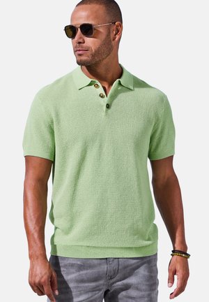 Mann trägt hellgrünes kurzärmeliges Strick-Poloshirt, graue Jeans, Sonnenbrille und Armbänder, schaut seitlich vor einfarbigem Hintergrund.