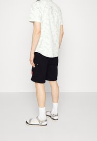 Camisa de manga corta con un patrón ligero, pantalones cortos negros con bolsillo lateral, calcetines blancos y sandalias con correas ajustables y una marca.