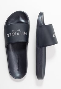 Mörkblåa slip-ons i en slät textur, med en bred rem och präglad "HILFIGER EST. 1985"-logotyp. Platt sula, avspänt design.