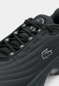 Μαύρο αθλητικό παπούτσι με υφή στο επάνω μέρος, διαθέτει σχέδιο από δίχτυ και ελαστικά κορδόνια. Το λογότυπο της Lacoste εμφανίζεται στο πλάι.