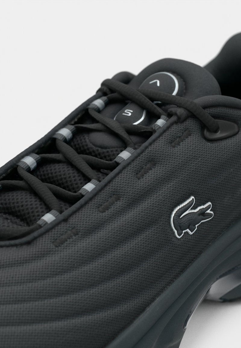 Sort atletiksko med en struktureret overdel, der har et mesh-design og elastiske snørebånd. Lacoste-logo vises på siden.