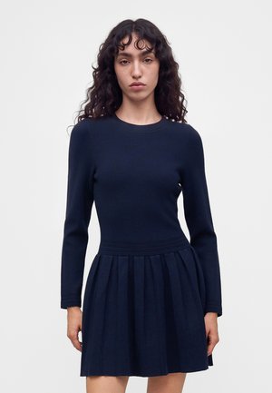 Jeune femme aux longs cheveux bouclés portant une robe bleu marine ajustée à manches longues avec une jupe plissée, debout devant un fond uni.