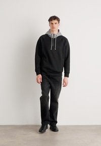 Sweatshirt negru cu glugă gri, șnururi și buzunar frontal, purtat cu blugi închiși la culoare și adidași negri. Modelul stă în fața unui fundal simplu.