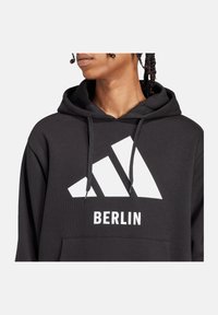 Felpa nera con cappuccio, caratterizzata da un grande logo Adidas bianco e la scritta "BERLIN". Presenta una tasca a marsupio e un cappuccio con cordino. Tessuto testurizzato.