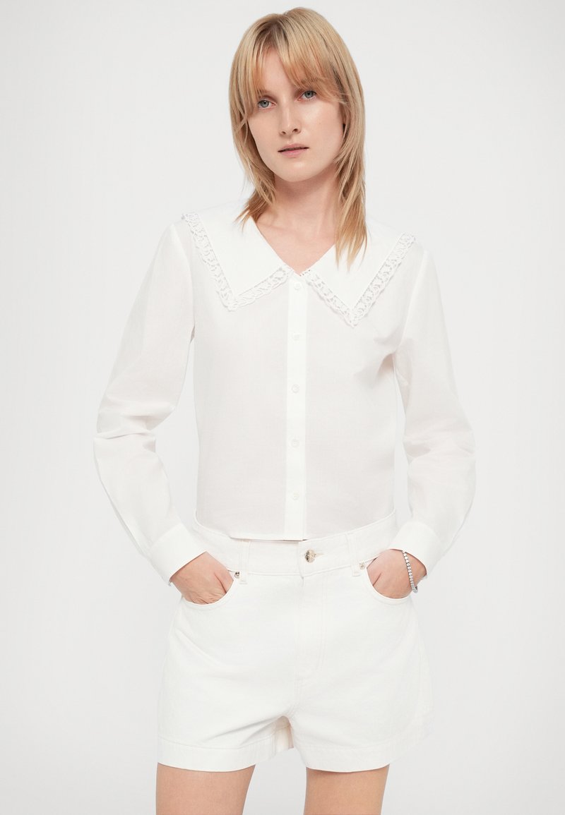 Chemise blanche à boutons avec un col bordé de dentelle, manches longues et coupe courte. Portée avec un short en denim blanc, les mains dans les poches.