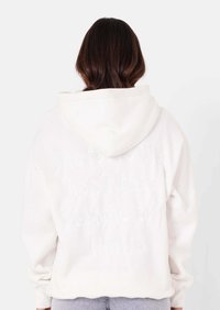 Persoon met lang donker haar draagt een witte hoodie met vaag reliëftekst op de achterkant, gecombineerd met lichtblauwe spijkerbroek.