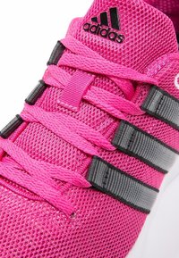 Zapatilla deportiva rosa con parte superior de malla, que presenta rayas y cordones negros, una superficie texturizada y un pequeño logotipo negro en la lengüeta.