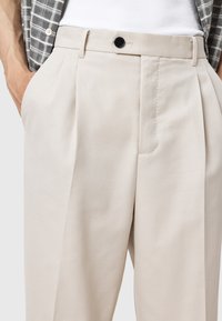 Beige pleated trousers with a smooth texture, featuring a front button closure and side pockets, displayed against a neutral background.