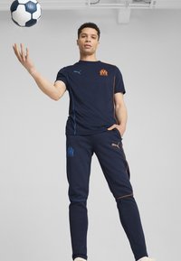 Jūrinės sportinės aprangos komplektas, kuriame yra trumpomis rankovėmis marškiniai su oranžiniais ir mėlynais akcentais, bei derančios kelnės su logotipo detalėmis; komplekte taip pat yra futbolo kamuolys.