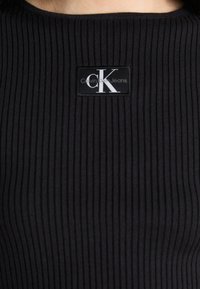 Melns ribots adījums tops ar noapaļotu kakla izgriezumu un taisnstūra etiķeti ar "Calvin Klein Jeans" logotipu baltā un pelēkā krāsā.