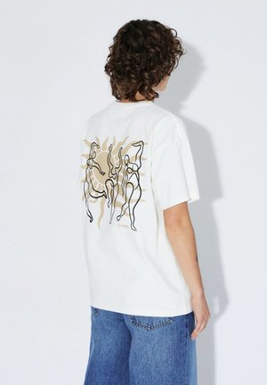 Persona con capelli ricci che indossa una maglietta bianca con un motivo astratto di sole e figure danzanti sulla schiena, abbinata a jeans blu.