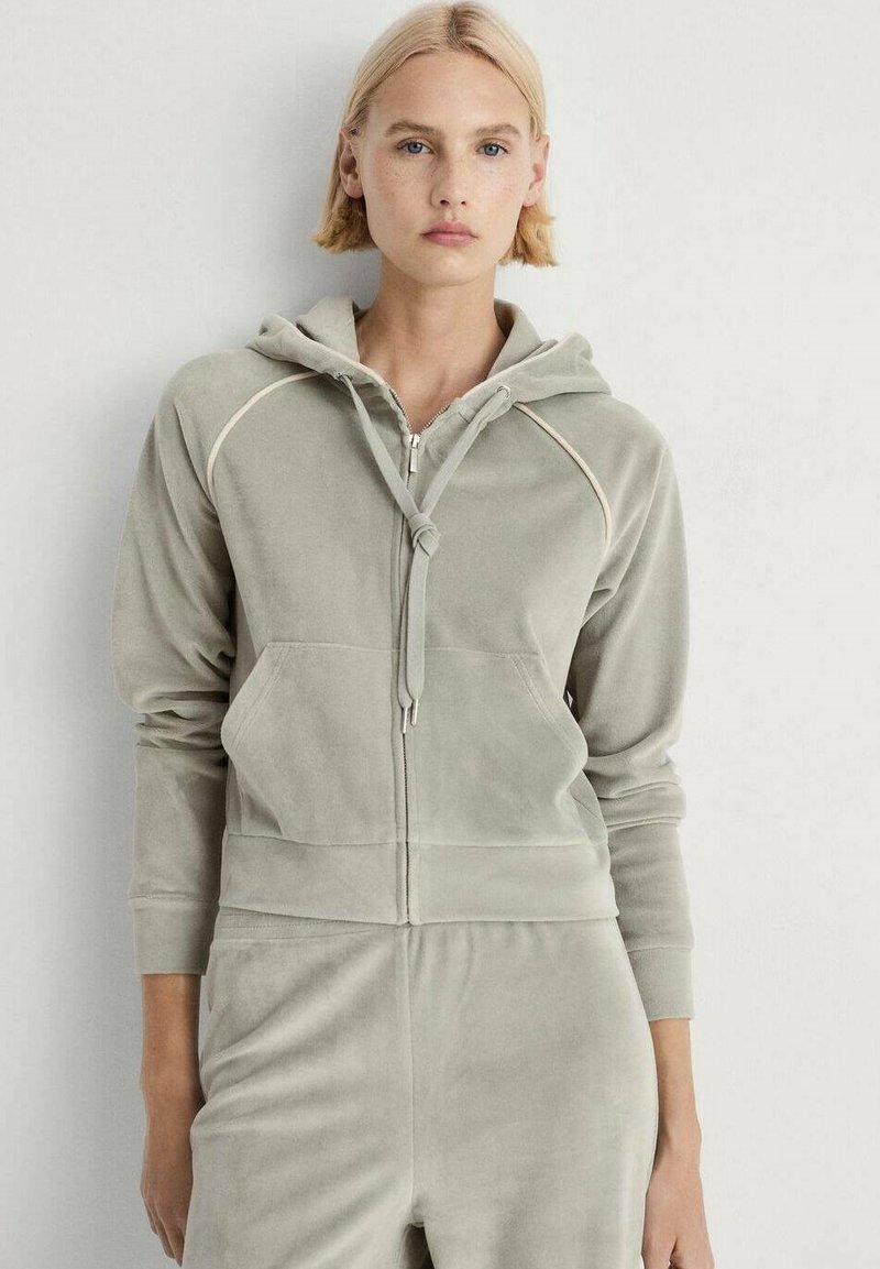 Felpa zip grigia chiara in morbido materiale vellutato. Presenta un cappuccio, polsini a costine e tasche davanti. Cuciture decorative lungo le giunture.