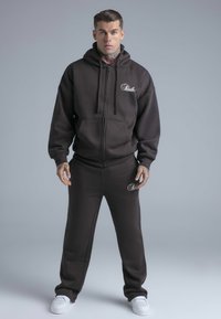 Bruine hoodie met rits, logo en voorzakken, gecombineerd met bijpassende joggingbroek. De stof is zacht en gestructureerd; heeft een casual pasvorm en wordt gedragen met witte sneakers.