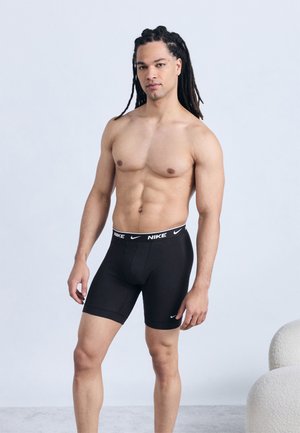 Άνδρας χωρίς πουκάμισο με μακριά πλεξούδα φοράει μαύρα εσώρουχα τύπου boxer της Nike, στέκεται μπροστά σε απλό φωτεινό φόντο.