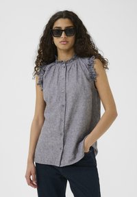 Halli hallituspluus, millel on volangkrae ja volangvarrukad, nööbitud esiosa ja tekstureeritud kangas. Kanda tume teksadega.