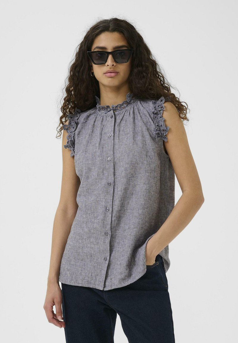 Halli hallituspluus, millel on volangkrae ja volangvarrukad, nööbitud esiosa ja tekstureeritud kangas. Kanda tume teksadega.