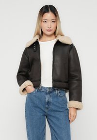 Joven mujer vestida con una chaqueta de borrego marrón oscuro sobre una camisa blanca y jeans de cintura alta azules, de pie con una mano en el bolsillo.