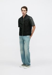 Juoda liemenė be rankovių su priekinio užtrauktuko uždarymu, dėvima ant pilkos trumpomis rankovėmis marškinėlių. Šviesiai mėlyni džinsai ir žali sportbačiai užbaigia aprangą.