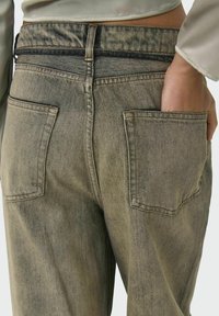 Farkut vaaleanbeige sävyssä, joissa on korkea vyötärö, takataskut ja tekstuurintuntuinen pinta. Ompeleet näkyvät saumoissa.