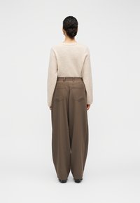 Μπεζ πλεκτό πουλόβερ με μακριά μανίκια, συνδυασμένο με καφέ παντελόνια ψηλόμεσα, με φαρδύ πόδι. Λείο ύφασμα, λιτή διακόσμηση και χωρίς ορατά μοτίβα.