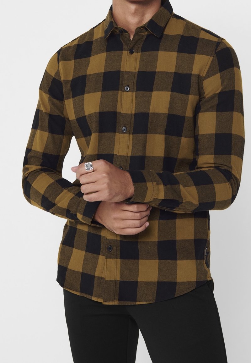 Knopenlange blouse in een geel en zwart ruitpatroon, gemaakt van zachte stof. Voorzien van een kraag, lange mouwen en zwarte knopen. Handen samengevoegd.