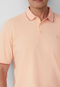 Mann trägt ein hellpfirsichfarbenes Poloshirt mit Kragen und kurzen Ärmeln, das ein kleines gesticktes Logo auf der Brust zeigt.