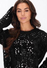 Czarny sweter z cekinami, o luźnym kroju, z błyszczącymi srebrnymi cekinami i miękkim, teksturowanym materiałem. Dekolt jest okrągły i nieco szeroki.
