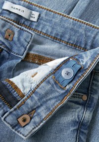 Detailní záběr modrých džín s kovovým zipem, knoflíkem, nastavitelným pásem v pase a viditelnou značkou "name it" uvnitř pasu.