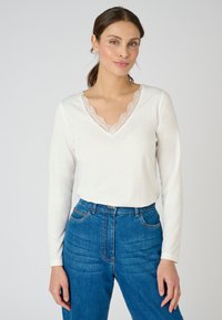 Witte langemouwen top met een V-hals en delicate kanten afwerking. Gecombineerd met hoog getailleerde donkerblauwe jeans met een gestructureerd patroon.