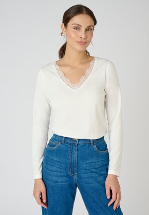 White long-sleeve top with a V-neck featuring delicate lace trim. Paired with high-waisted dark blue jeans with a textured pattern.
