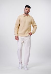 Stickad krämfärgad tröja med rundad krage, i kombination med vita byxor och vita sneakers. Slät textur och avslappnad passform.