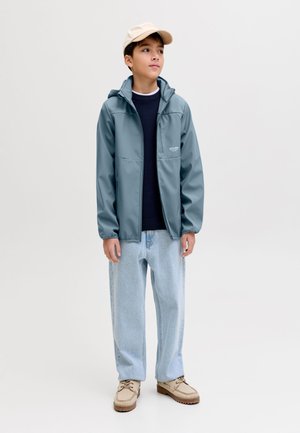 Αγόρι που στέκεται φορώντας μπλε μπουφάν με κουκούλα, μαύρο πουλόβερ, ανοιχτό μπλε τζιν, μπεζ παπούτσια και μπεζ καπέλο, κοιτάζει ελαφρώς στο πλάι.