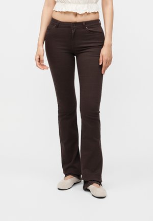 Femme portant un pantalon évasé taille haute marron foncé, un crop top blanc et des chaussures plates beiges, debout devant un fond clair et uni.