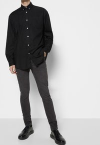 Hombre vestido con una camisa negra de botones, jeans ajustados gris oscuro y zapatos de cuero negros con cordones, de pie con una mano en el bolsillo.